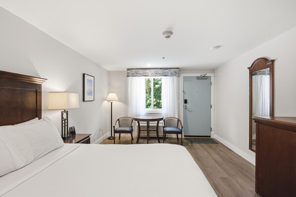 A hotel room with a bed, two chairs, a table, a desk, a mirror, and a door leading to another room.
