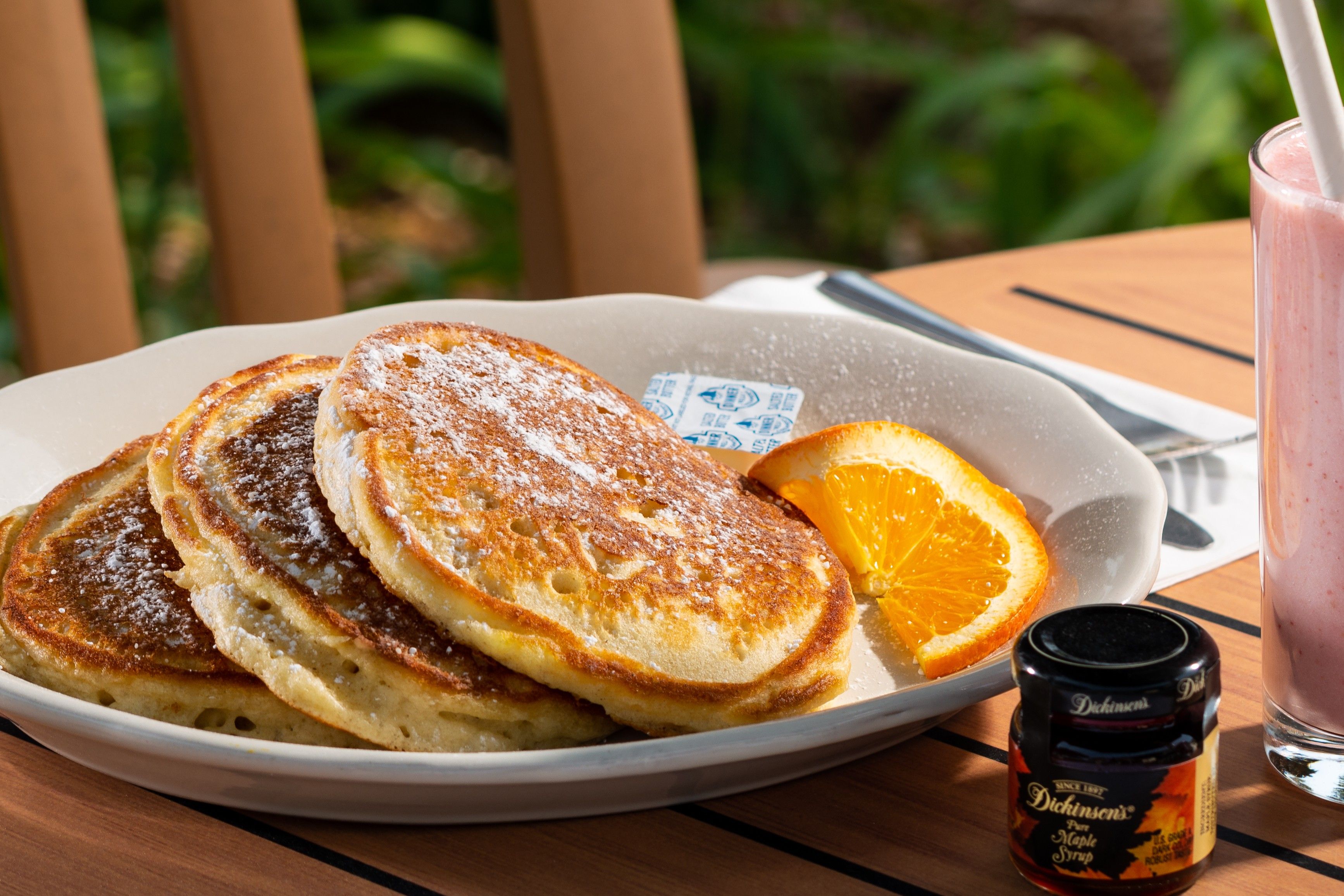 A plate of pancakes with powdered sugar, an orange slice, and a bottle of maple syrup on a table.