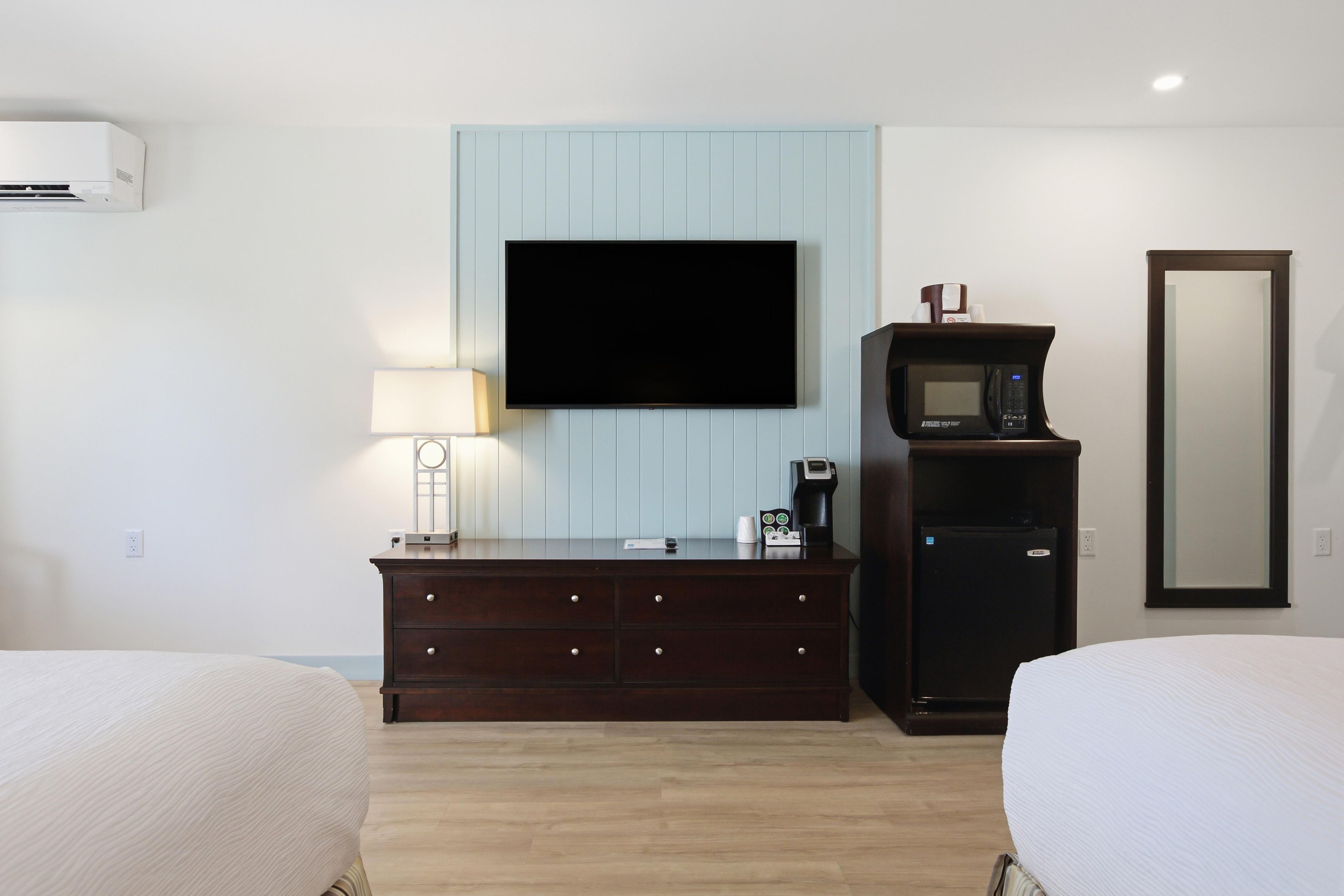 A hotel room with a flat-screen TV mounted on the wall, a dark wooden dresser with drawers, a coffee maker, and a microwave. There are two beds with white bedding.