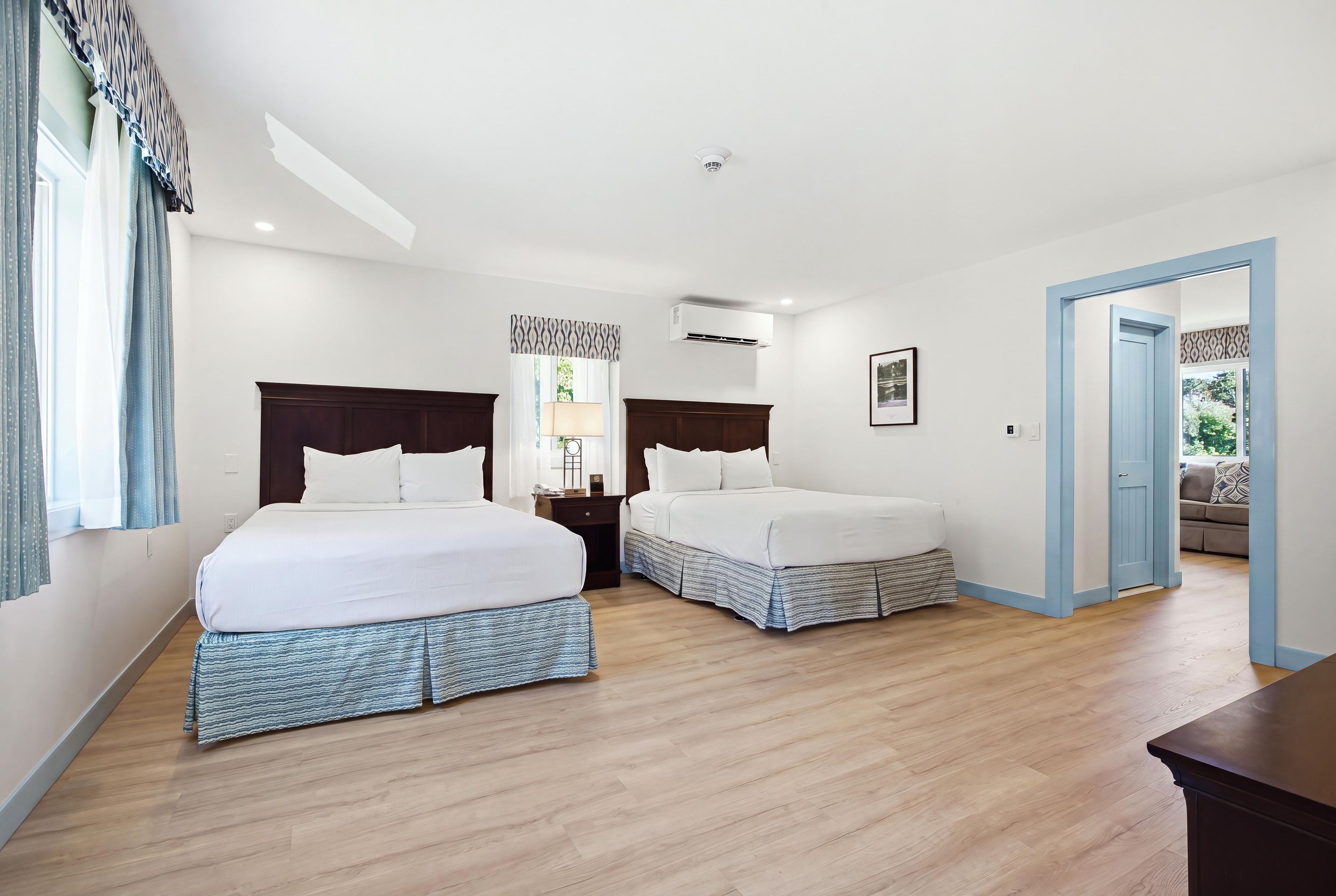 A hotel room with two beds, a nightstand, a lamp, an air conditioning unit, and a door leading to another room.