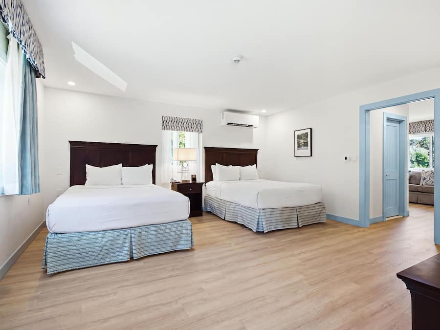 A hotel room with two beds, a nightstand, a lamp, an air conditioning unit, and a door leading to another room.