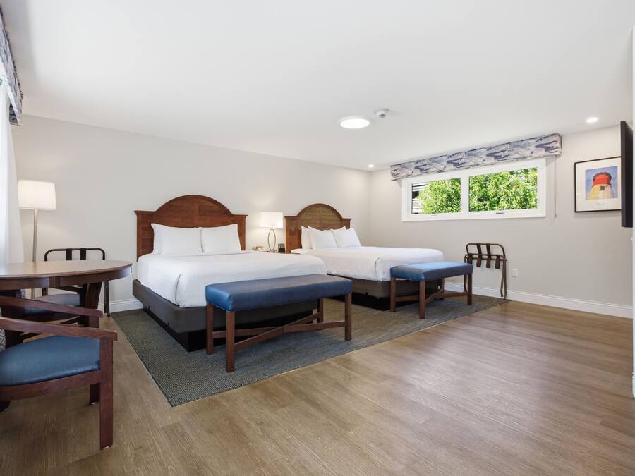 A hotel room with two beds, a desk, chairs, a window with a view, and a lamp.