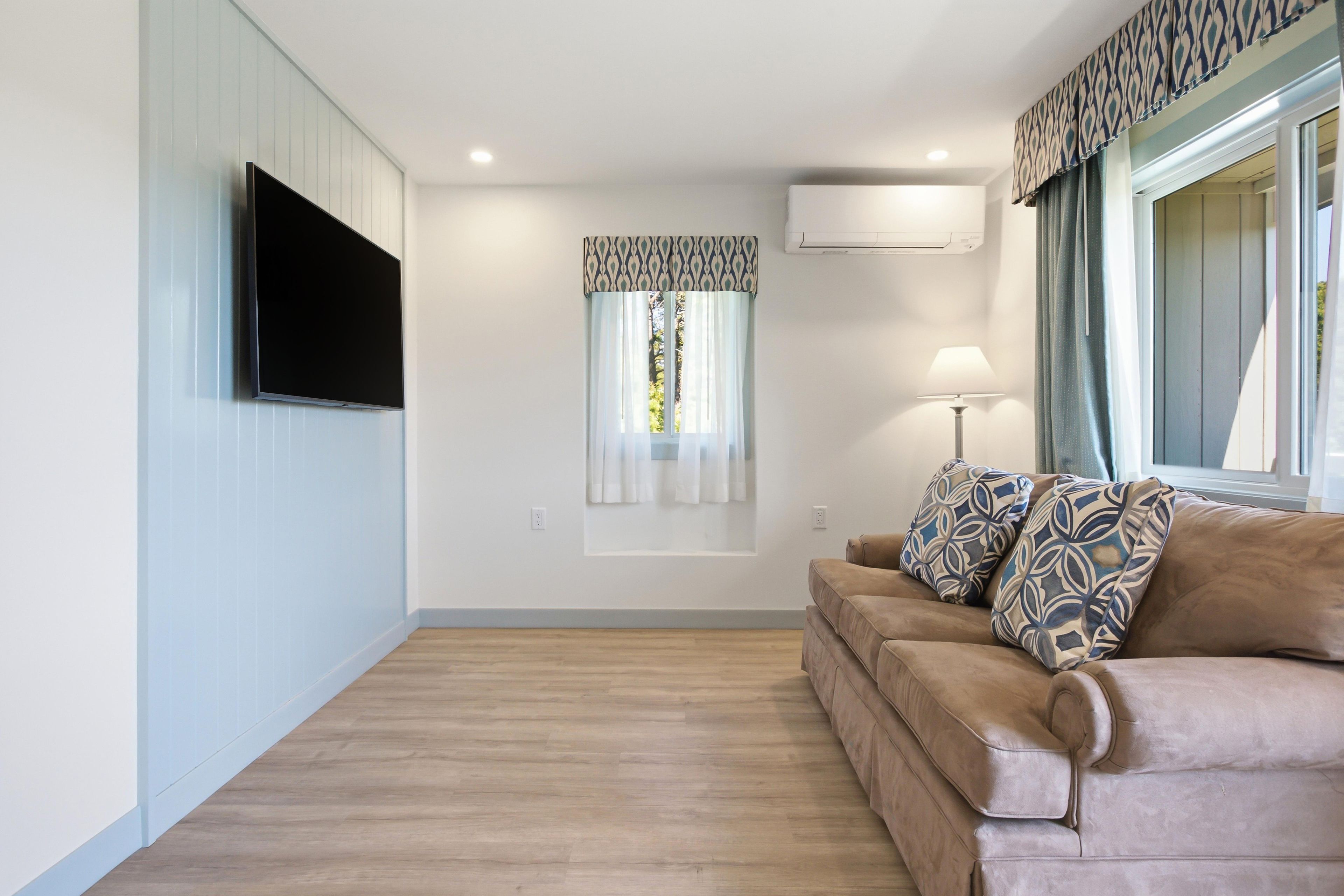 A living room with a flat-screen TV mounted on the wall, a window with patterned curtains, a sofa with patterned cushions, and an air conditioning unit on the wall.