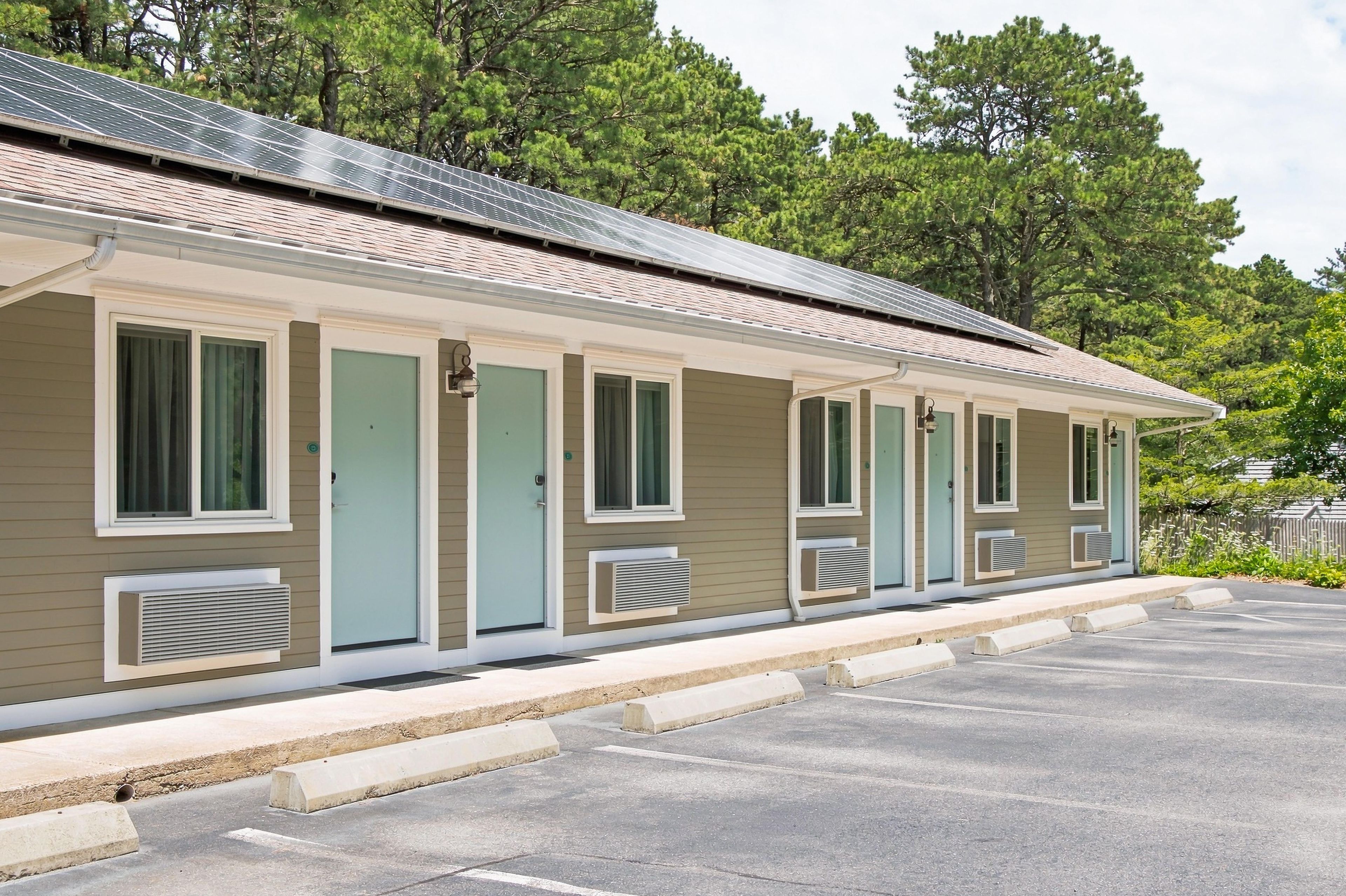 A row of motel rooms with individual air conditioning units, a covered parking area, and a forested backdrop.