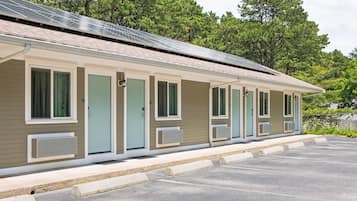 A row of motel rooms with individual air conditioning units, a covered parking area, and a forested backdrop.