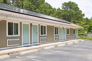 Filas de habitaciones de motel con unidades de aire acondicionado individuales, zona de estacionamiento techada y un paisaje boscoso de fondo.