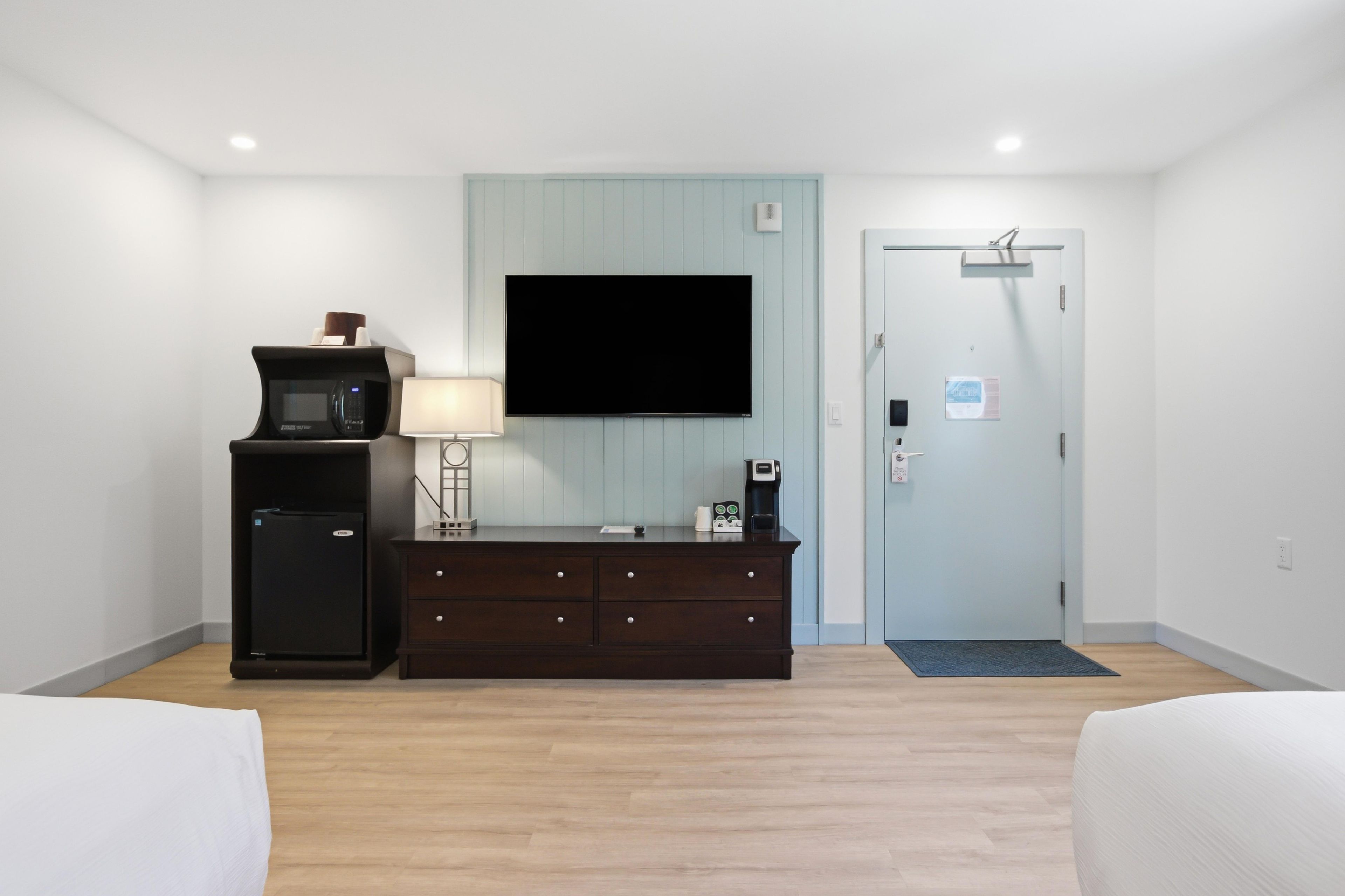A hotel room with a flat-screen TV mounted on the wall, a small refrigerator, a dark wooden dresser, a lamp, and a door with a sign.