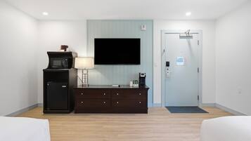A hotel room with a flat-screen TV mounted on the wall, a small refrigerator, a dark wooden dresser, a lamp, and a door with a sign.