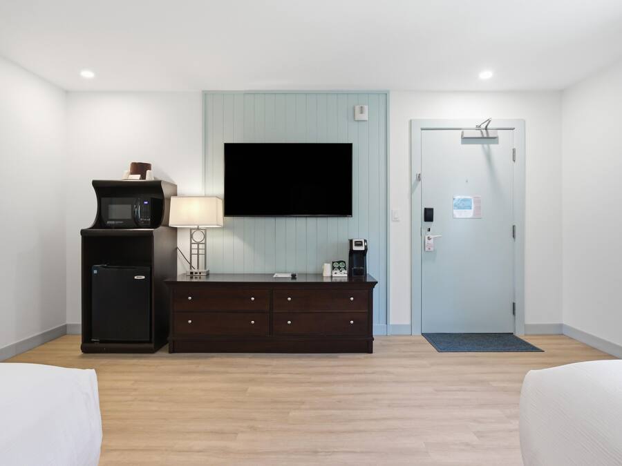 Quarto de hotel com televisão de tela plana na parede, minibar, cômoda de madeira escura, luminária e porta com placa.