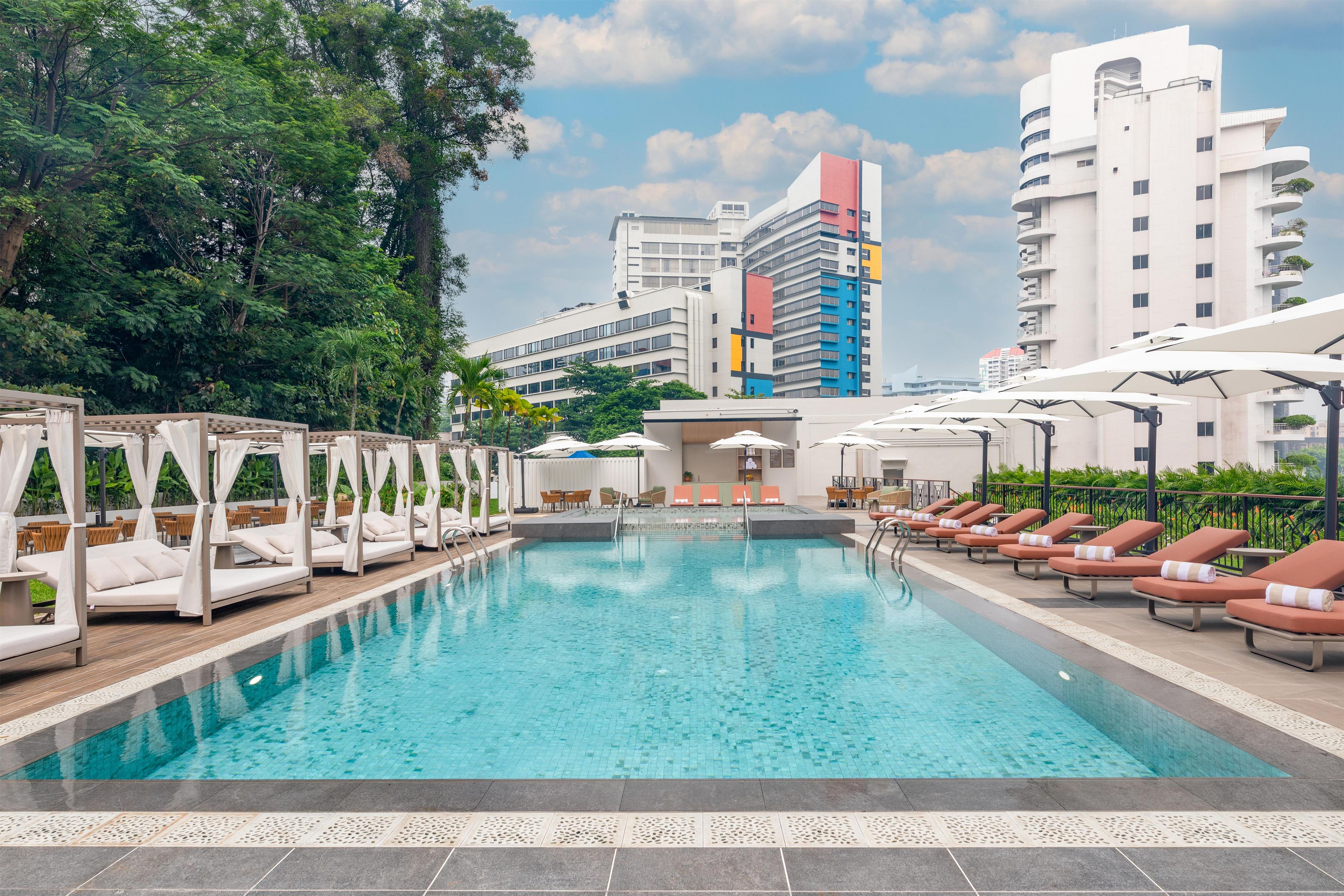 A swimming pool with lounge chairs and a canopy structure, surrounded by greenery and modern buildings.