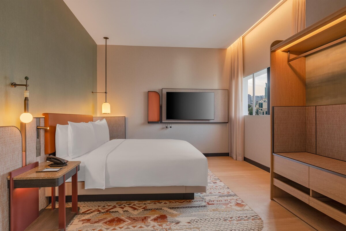 Habitación de hotel con cama, mesita de noche, lámpara, televisor y vista a la ciudad.