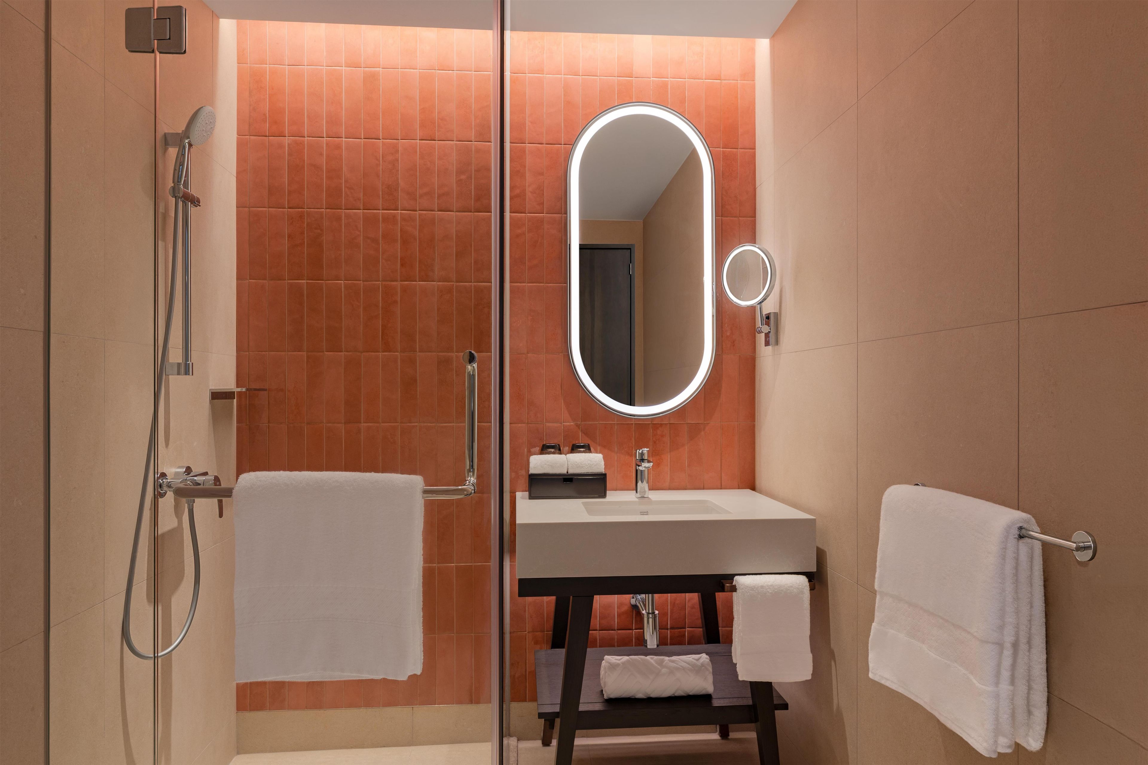 A modern bathroom with a glass shower enclosure, a round mirror with integrated lighting, and a dark wooden vanity with a white sink.