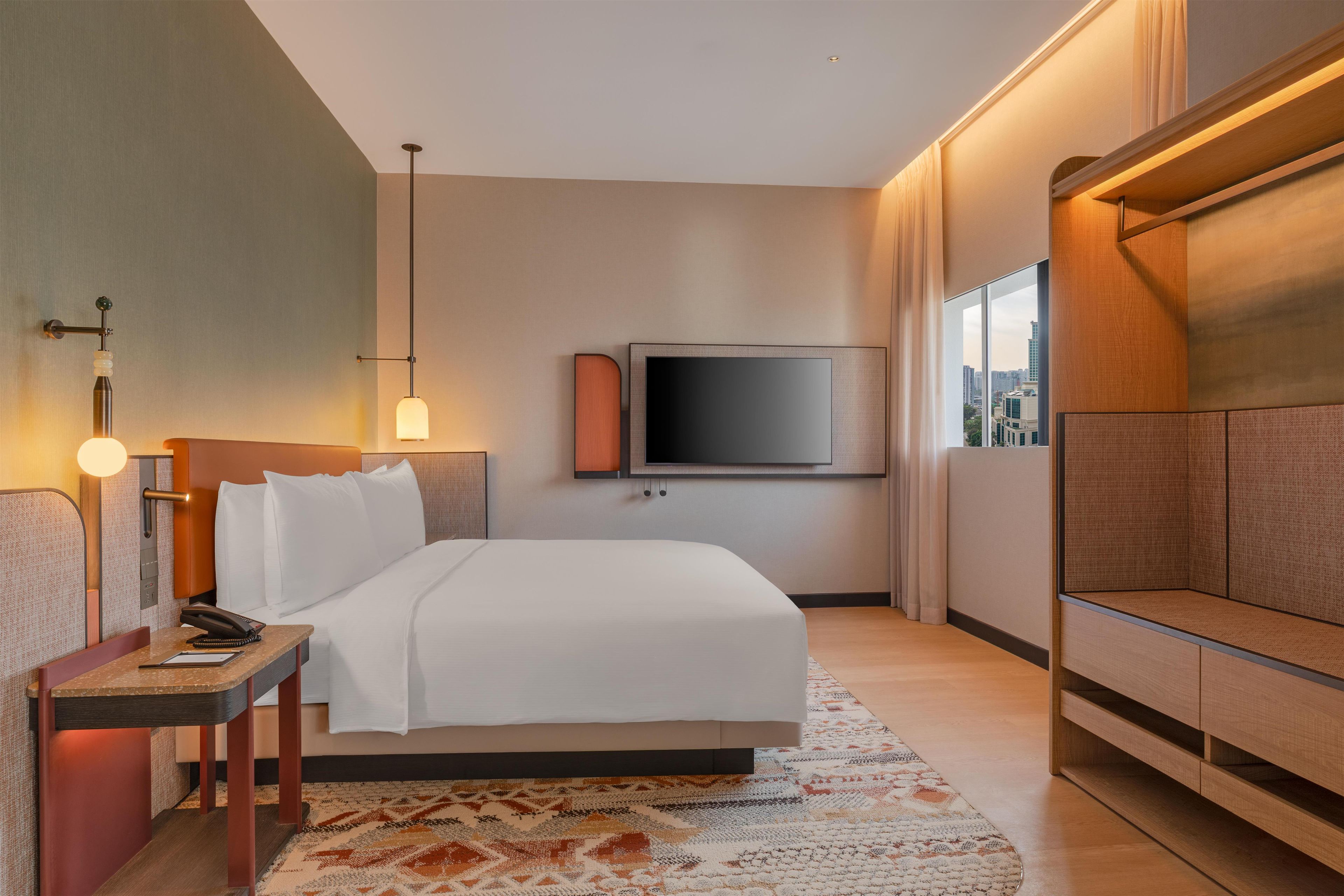 A hotel room with a bed, bedside table, lamp, TV, and a view of the city.