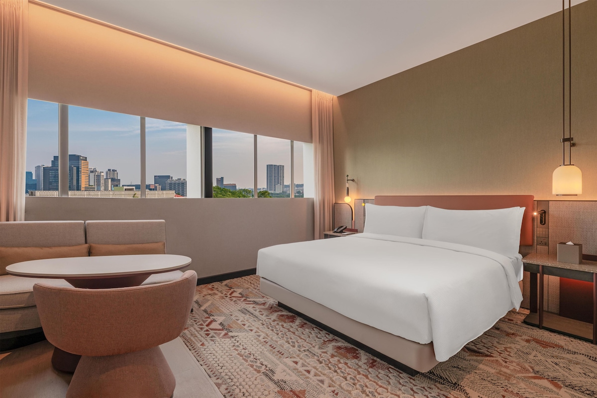 A hotel room with a bed, a sofa, a small table, a nightstand, and a view of the city skyline through large windows.