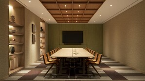 Un salón de conferencias con una larga mesa, sillas, una pantalla de proyector y estantes con libros y objetos decorativos.