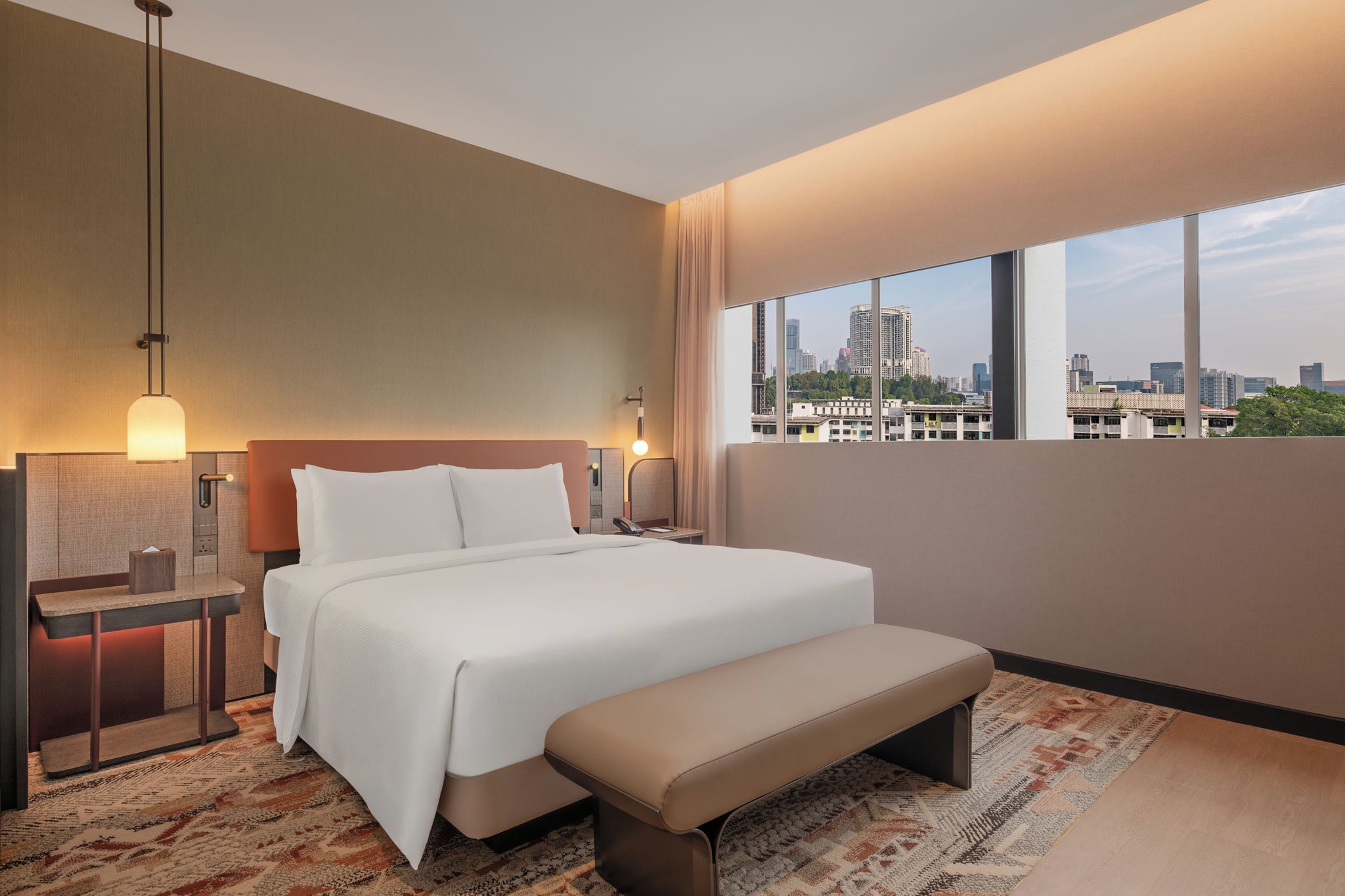 Une chambre d’hôtel moderne avec un grand lit, une banquette, une table de chevet et une vue sur la ville.