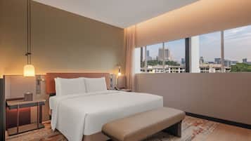 A modern hotel room with a large bed, a bench, a nightstand, and a city view.