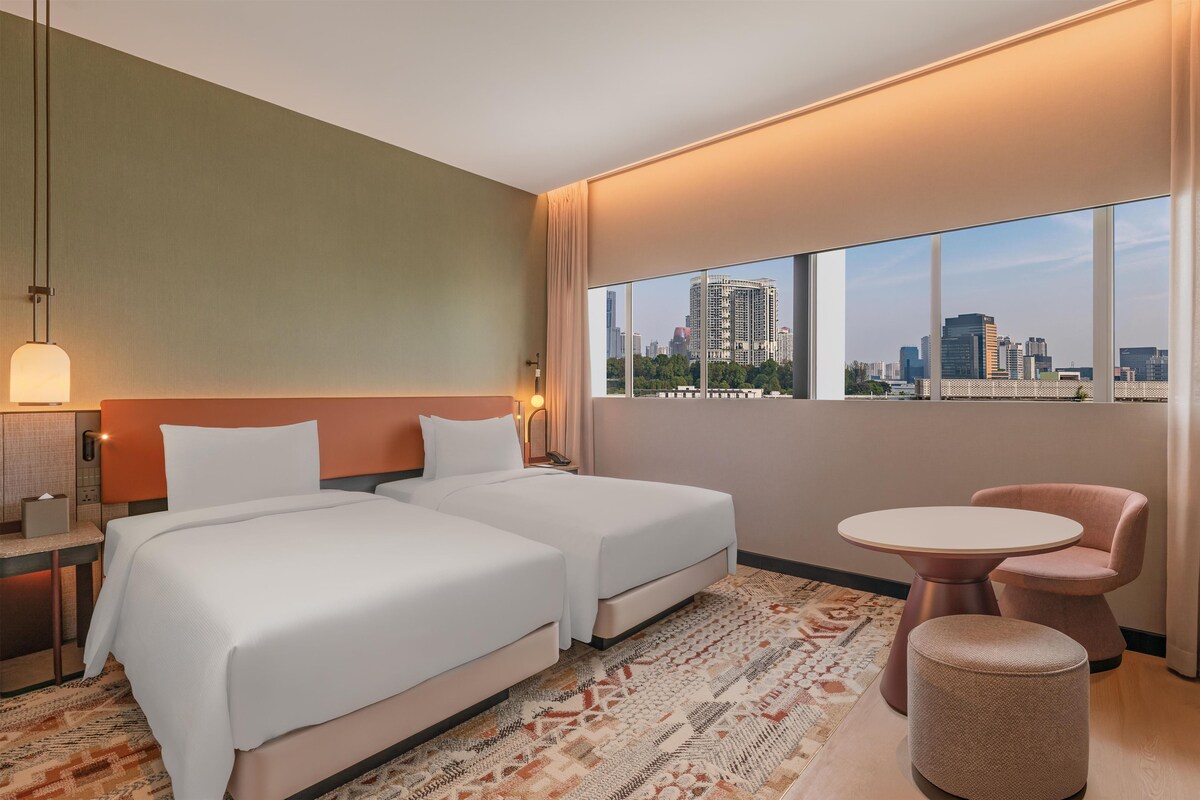 A hotel room with two beds, a small table, a chair, and a view of the cityscape.