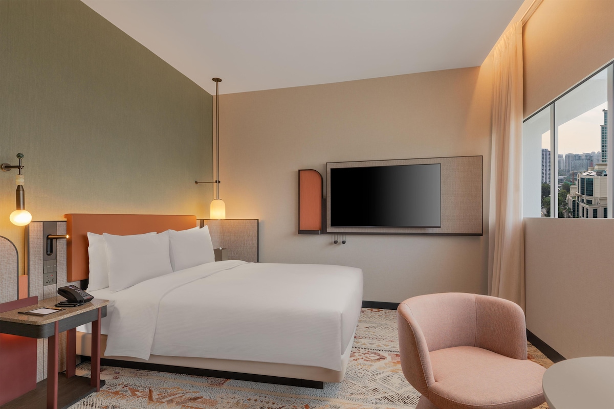 A hotel room with a bed, bedside table, lamp, TV, chair, and a view of the city.