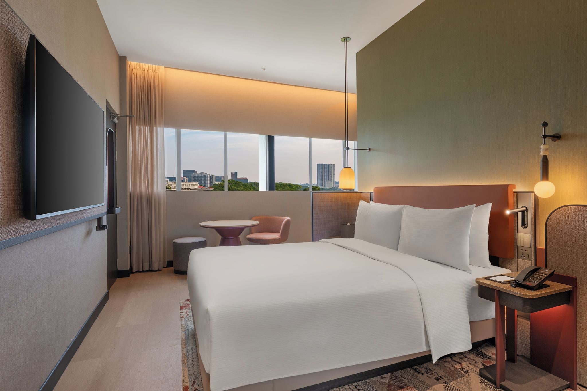 Habitación de hotel con una cama grande, un escritorio con teléfono, una silla, una mesita, un televisor en la pared y una ventana con vista a la ciudad.