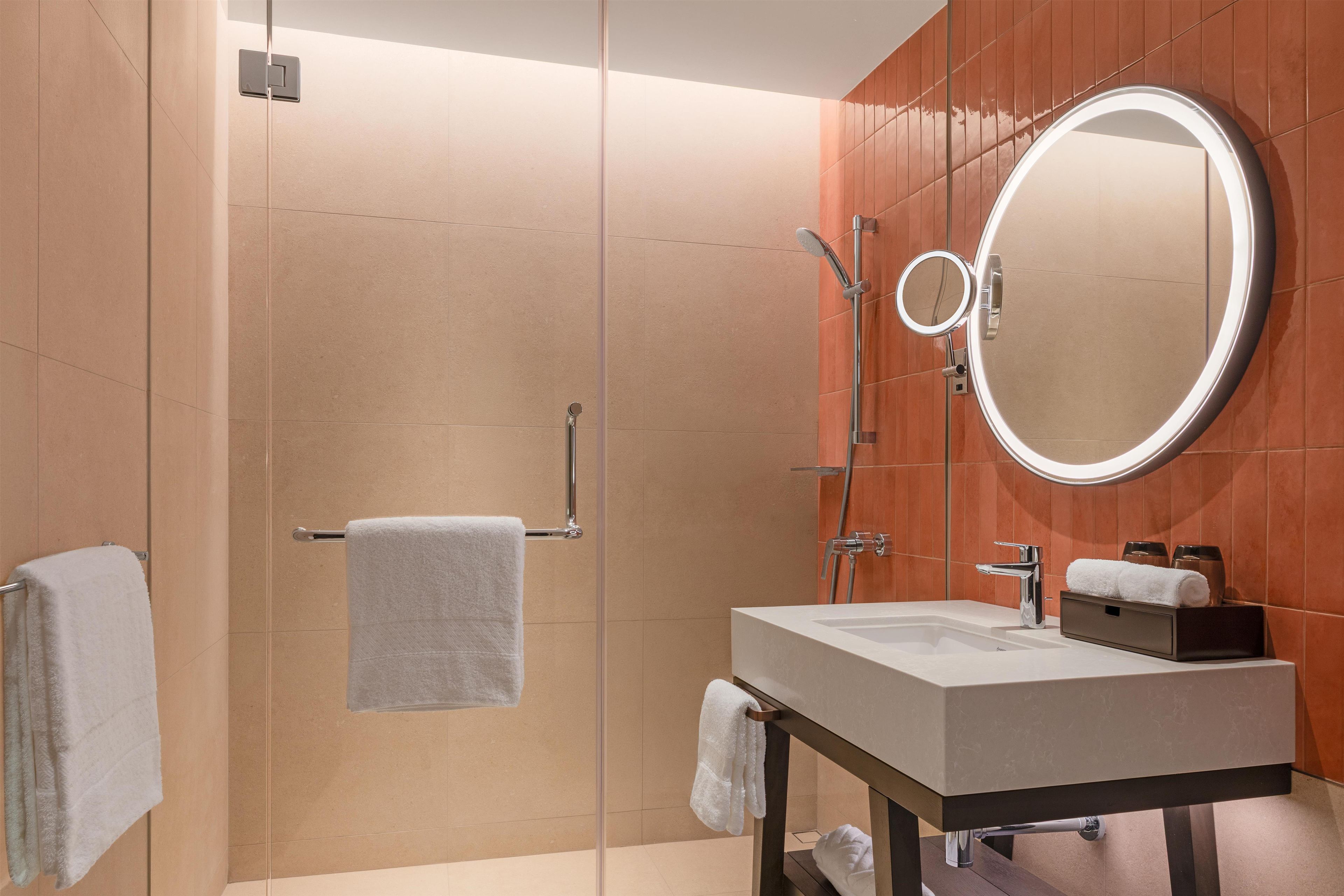A modern bathroom with a glass shower enclosure, a round mirror, a sink, and a towel rack.