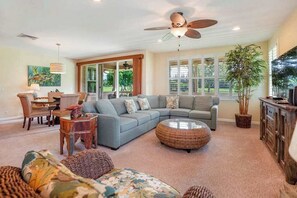 Ein Wohnzimmer mit einem Ecksofa, einem runden Kaffeetisch aus Rattan, einem Deckenventilator und einem großen Fenster mit Jalousien.