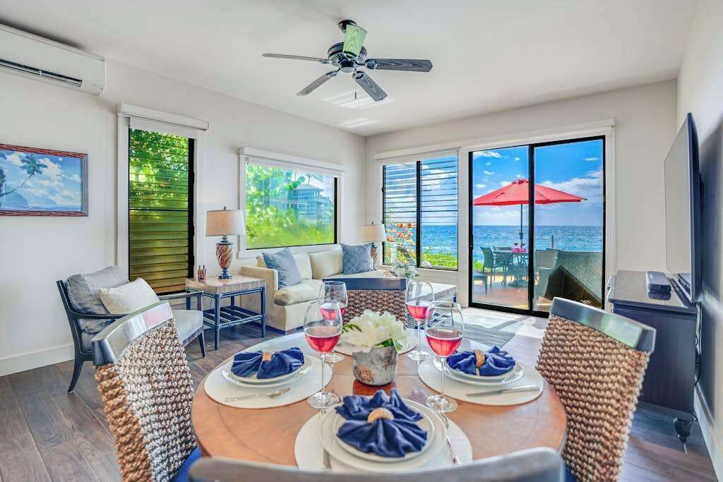 A dining area with a table set for four, a ceiling fan, and large windows with a view of the ocean.