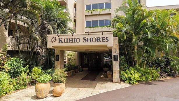 En hotellentrén med en skylt som visar texten ”KUHIO SHORES”, omgiven av tropiska växter och en gångbana med plattor.