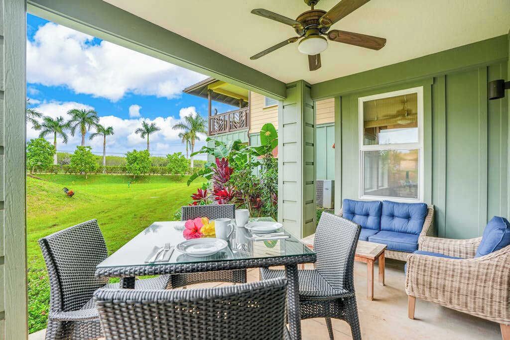 A room with a glass table, wicker chairs, a ceiling fan, and a view of a green lawn with palm trees.