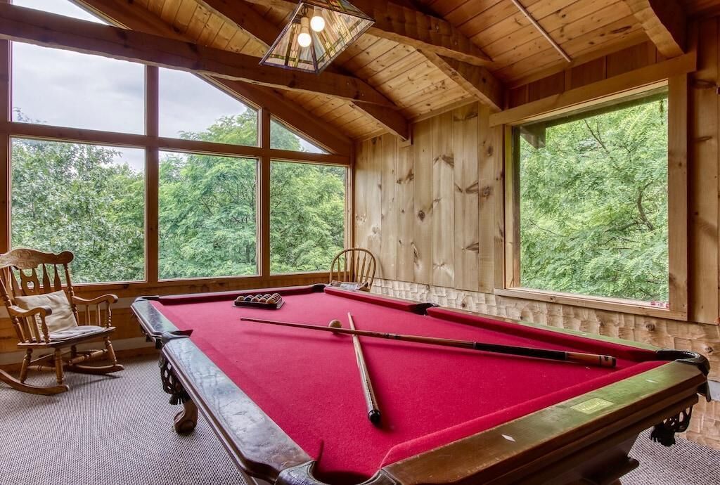 A pool table with a red felt surface, wooden walls, and large windows offering a view of the forest.