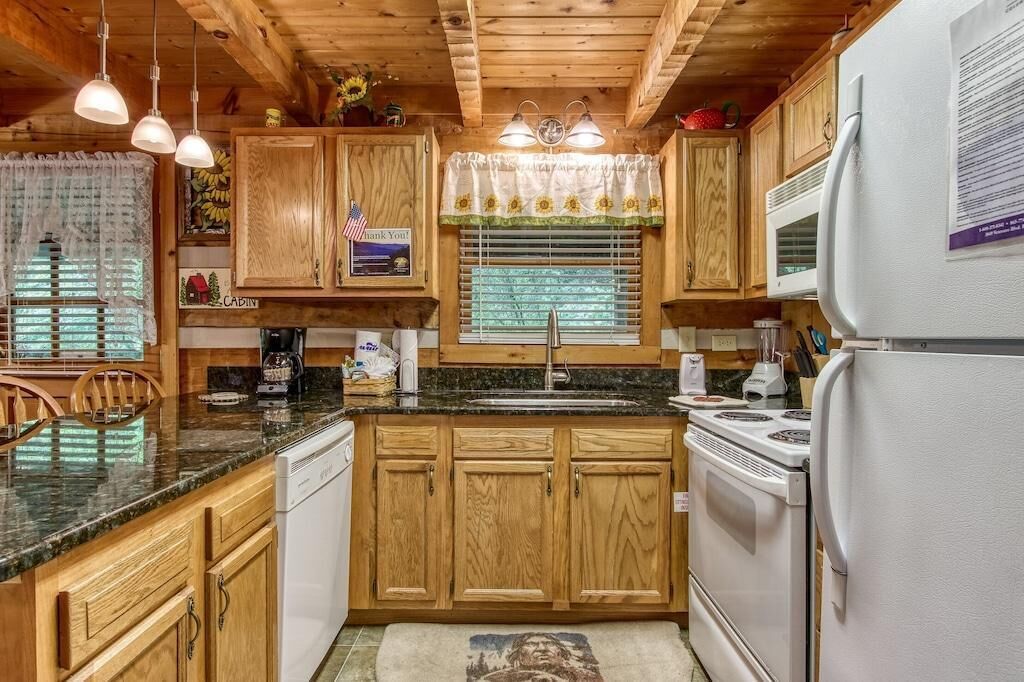 A cabin-style kitchen with wooden cabinets, a granite countertop, a stove, a refrigerator, and a window with a sunflower curtain.