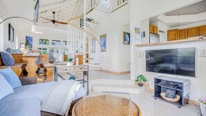 A spacious living area with a large TV, a round glass table, a staircase, and wooden furniture. - Princeville Paniolo 202A/202B Duplex- AC, Pool, Laundry- Hanalei Mountain Views (Princeville)