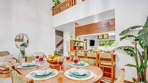 Un comedor con una mesa redonda preparada para cuatro personas, una cocina al fondo y una planta grande a la derecha.