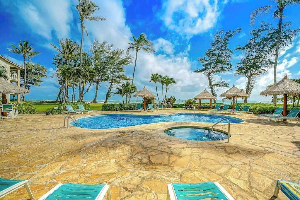 Una piscina con sillas de descanso, palmeras y una estructura de techo de paja.