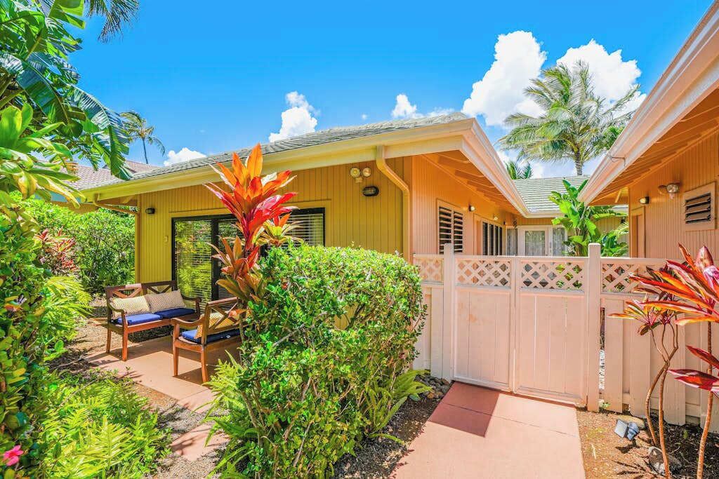 A yellow building with a covered patio, a wooden fence, and lush greenery.