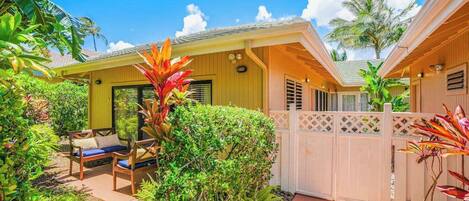 A yellow building with a covered patio, a wooden fence, and lush greenery.