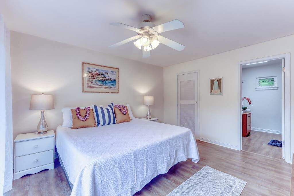 Ein Schlafzimmer mit einem Bett, einem Deckenventilator, einem Nachttisch, einer Lampe, einem gerahmten Bild an der Wand und einer Tür zu einem anderen Raum.