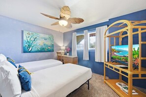 Ein Hotelzimmer mit zwei Betten, einem Deckenventilator, einem Gemälde, einem Schreibtisch mit Lampe, einem Fernseher auf einem Podest und einem Fenster mit Vorhängen.