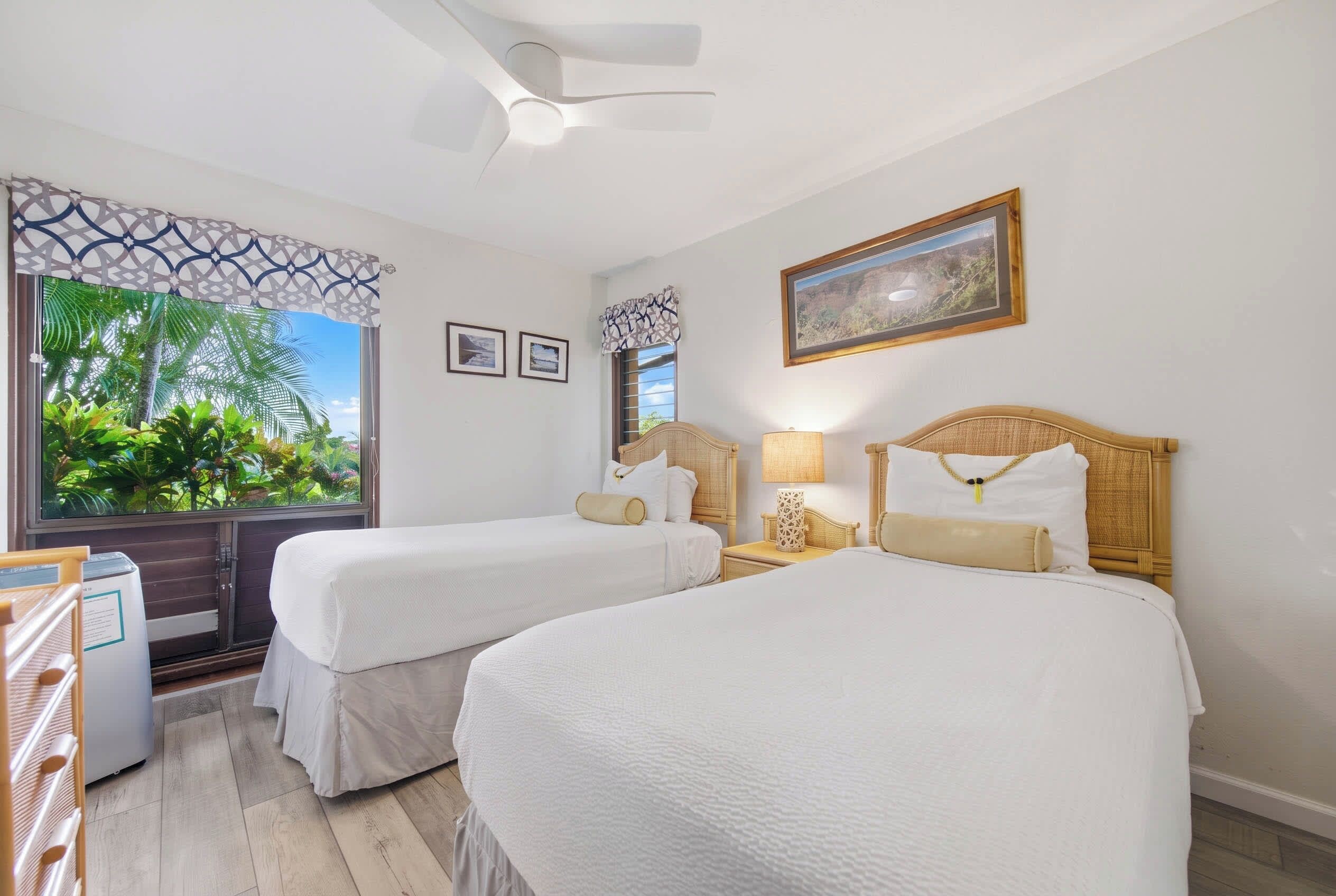 A hotel room with two beds, a desk, a window with a view of greenery, and a ceiling fan.