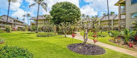 Un patio de hotel con césped cuidado, palmeras y un jardín circular central con un árbol y flores rojas.