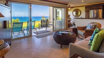 A spacious living area with a large sliding glass door leading to a balcony with a view of the ocean. There is a round wicker coffee table, a sofa with cushions, and a dining area with chairs.