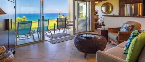 A spacious living area with a large sliding glass door leading to a balcony with a view of the ocean. There is a round wicker coffee table, a sofa with cushions, and a dining area with chairs.