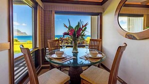 A dining area with a glass table, wooden chairs, a vase with flowers, and a view of the ocean. - New Bali Hai and Hanalei Bay Views from Princeville Bluff (Princeville)