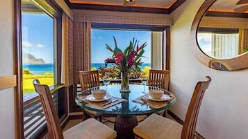 A dining area with a glass table, wooden chairs, a vase with flowers, and a view of the ocean.