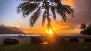 A sunset over a beach with a palm tree, rocks, and a distant mountain range. - NEW Fall $ale Walk to Hanalei Bay from a Waterfall View Home TVNC#5120 (Hanalei)