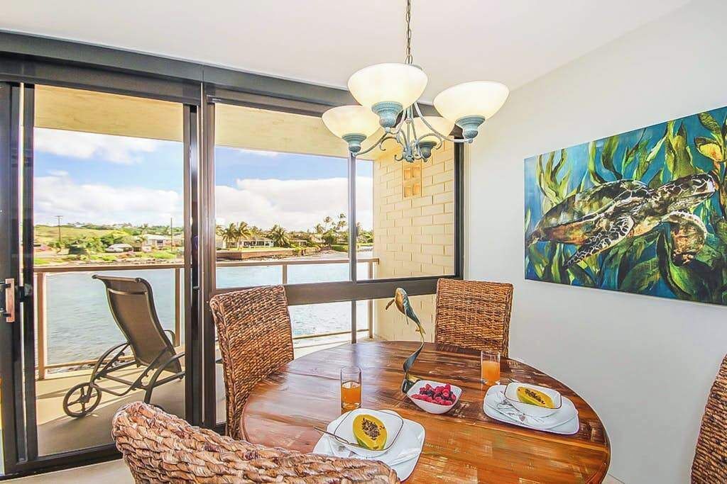 A dining area with a view of the water, wicker chairs, a round table, and a painting of a sea turtle.