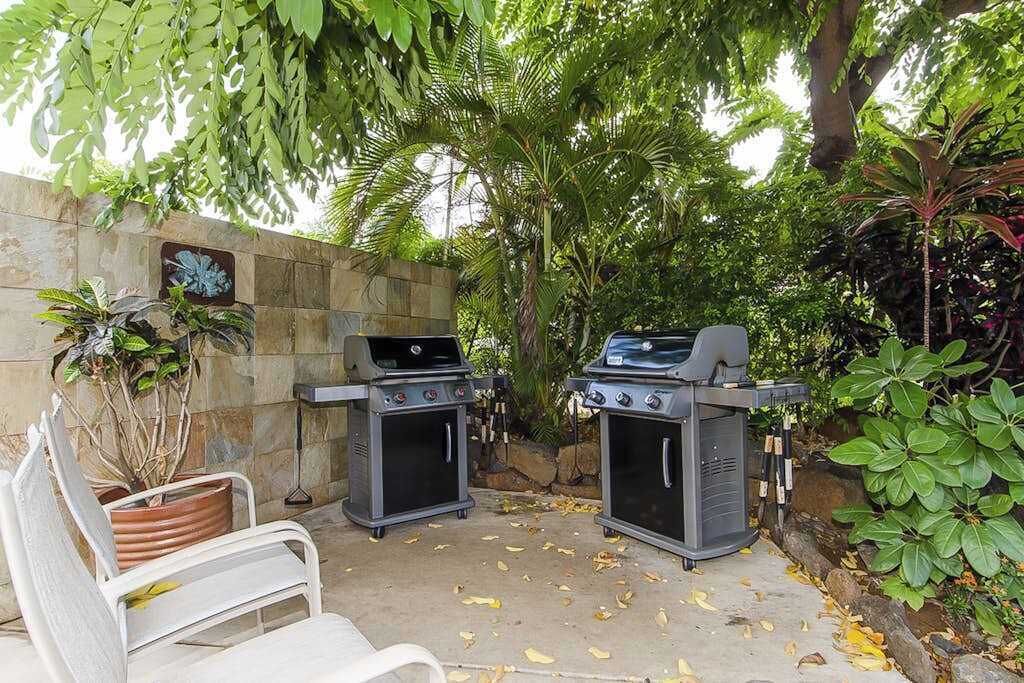 An outdoor patio with two barbecue grills, a white metal bench, and a potted plant.