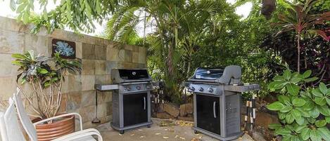 An outdoor patio with two barbecue grills, a white metal bench, and a potted plant.