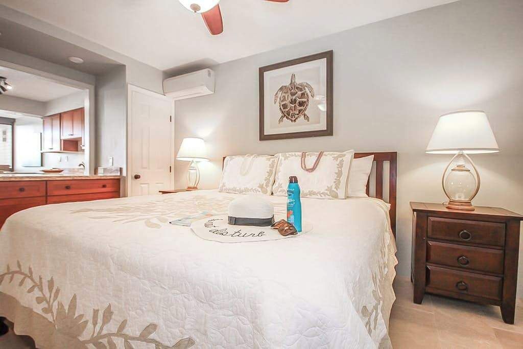A bedroom with a bed, bedside lamps, a nightstand, a framed picture on the wall, and a view of the kitchen.