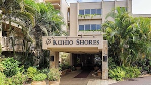 Hotellin sisäänkäynti, jonka yläpuolella on kyltti, jossa lukee ”KUHIO SHORES”. Sisäänkäyntiä ympäröi vehreä kasvillisuus ja palmuja.